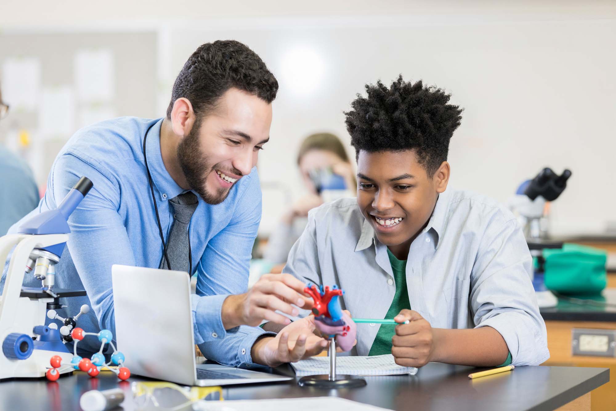 A teenage boy sits at a table in his High School science lab and studies a model of the human heart.  He attentive male teacher leans on the table and manipulates the model to answer his questions.